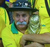 Old man with a mustache hugging a world cup while watching Brasil lose
